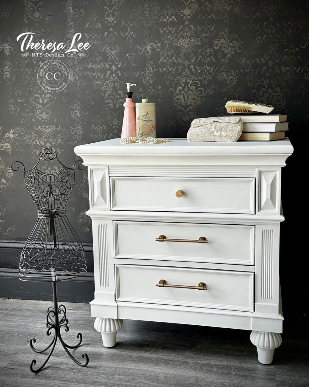 A white vintage nightstand with gold handles stands against a dark, ornate wallpaper. On top are books, a purse, and toiletries. A decorative black wire mannequin stands to the left. "Theresa Lee NTS Design Co." watermark is in the corner.