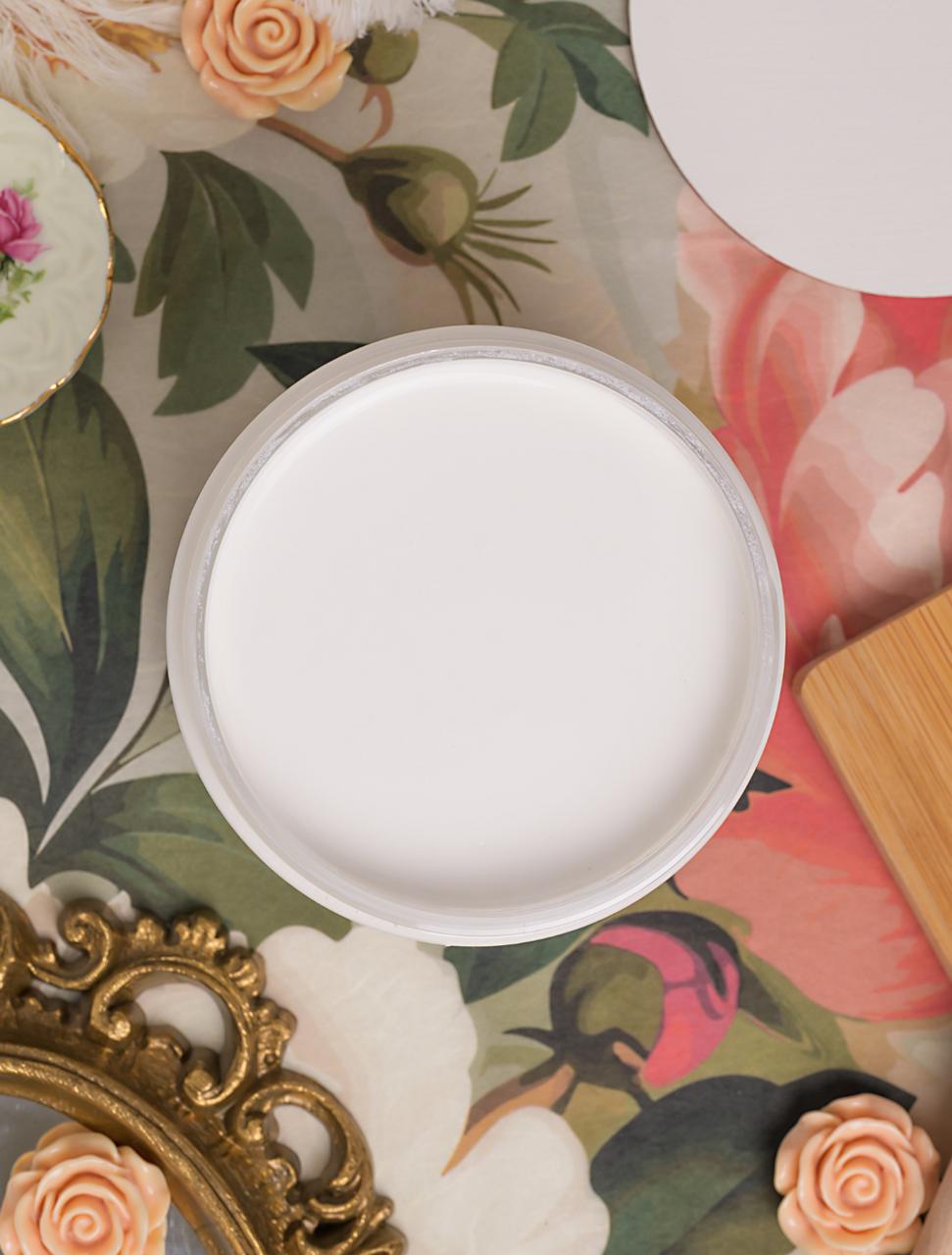 A round container filled with white paint sits on a floral-patterned surface, surrounded by decorative items including a gold frame, flowers, and a china saucer with a pink flower design.