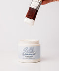 A hand holds a paintbrush with white paint dripping from it over an open container labeled "Silk All-In-One Mineral Paint, Elderflower." The background is plain white.