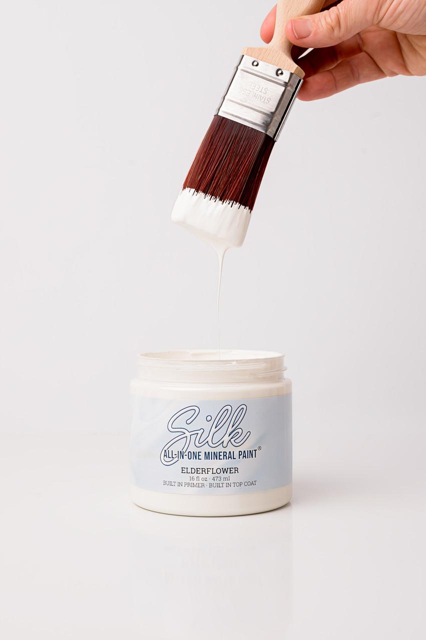 A hand holds a paintbrush with white paint dripping from it over an open container labeled "Silk All-In-One Mineral Paint, Elderflower." The background is plain white.