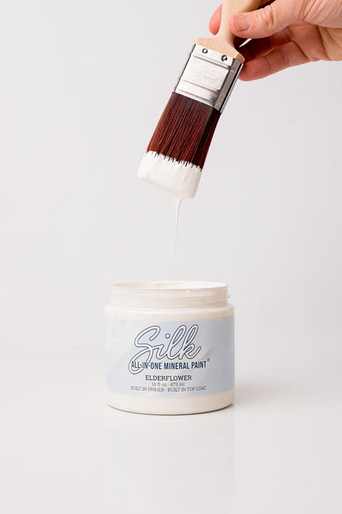 A hand holds a paintbrush with white paint dripping from it over an open container labeled "Silk All-In-One Mineral Paint, Elderflower." The background is plain white.