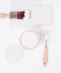A paintbrush with white paint, a round container of white paint, a palette knife, and a square and circle of white paint samples are arranged neatly on a white background.