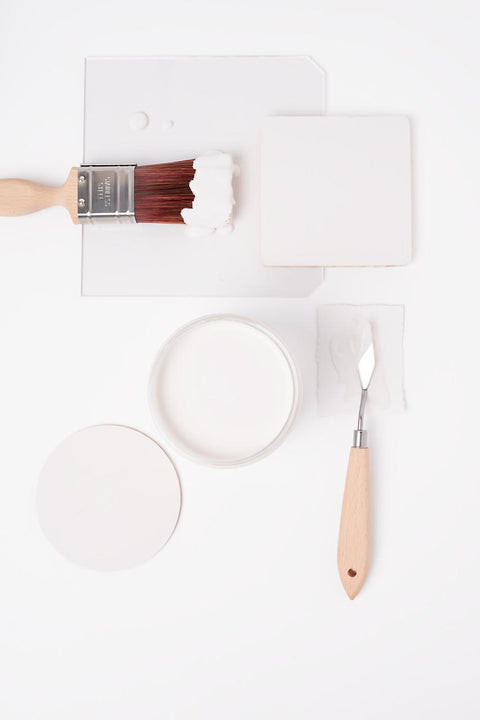 A paintbrush with white paint, a round container of white paint, a palette knife, and a square and circle of white paint samples are arranged neatly on a white background.