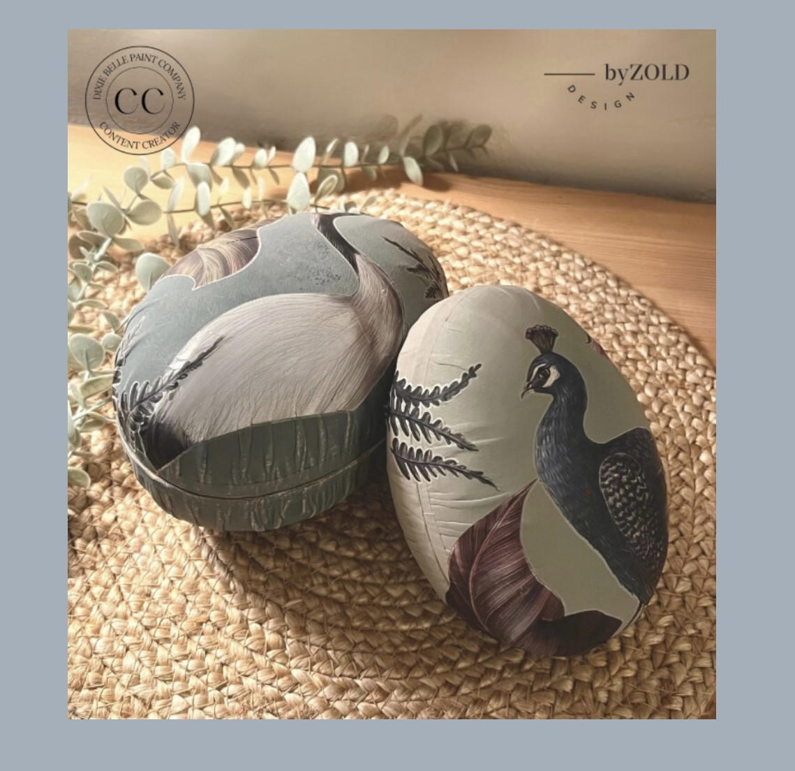Bluish teal painted faux eggs with the Elegant Avian Flora Transfer applied on them. Staged on a woven placemat next to faux eucalyptus.