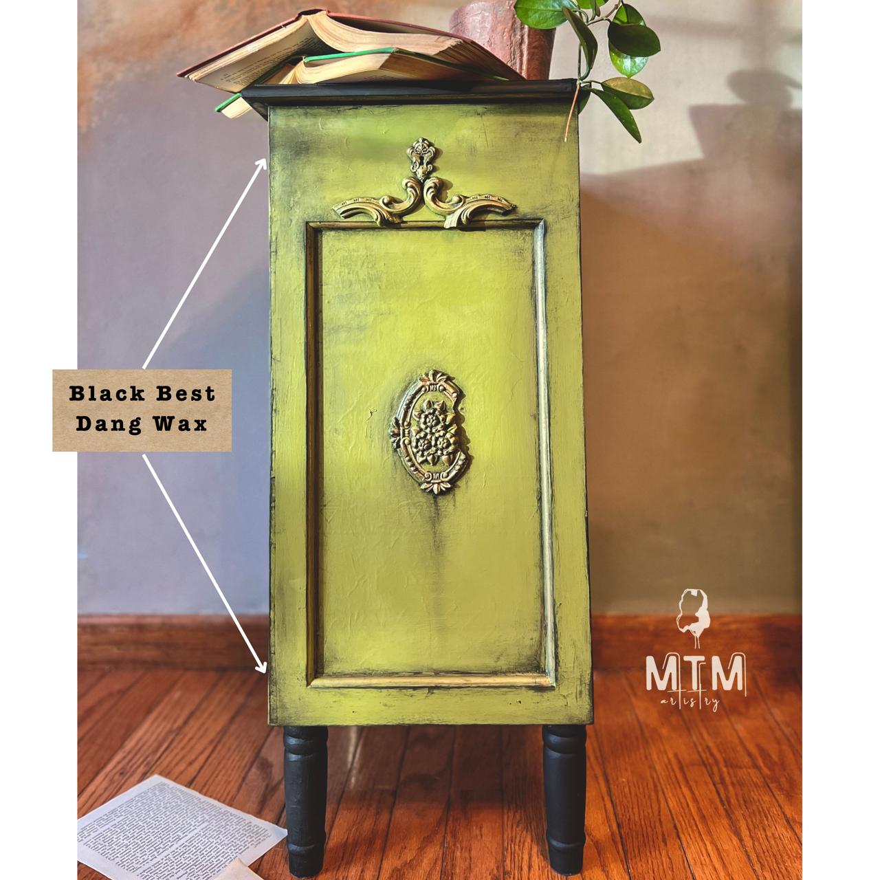 A small, ornate green cabinet with black wax accents stands on a wooden floor. An open book and a plant adorn the top. A paper is on the floor nearby. Text reads "Black Best Dang Wax" with arrows pointing to the cabinet.