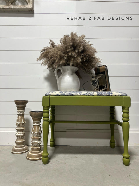 Green painted bench makeover with white and blue upholstery on the seat. Staged with an old camera and a vase of cattails on top. Two large candlesticks to the left of the bench.