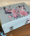 A light blue wooden box decorated with illustrations of cranes, pink peonies, and butterflies. It sits on a wooden surface with decorative beads and artificial flowers nearby. The box has ornate metal latches.