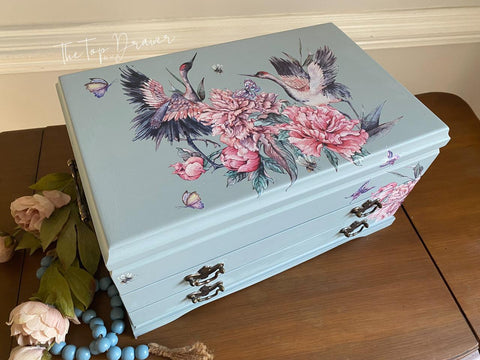 A light blue wooden box decorated with illustrations of cranes, pink peonies, and butterflies. It sits on a wooden surface with decorative beads and artificial flowers nearby. The box has ornate metal latches.