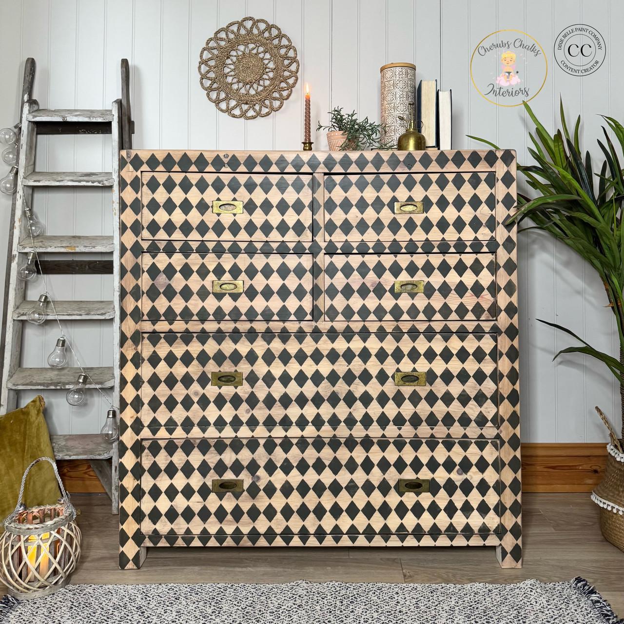 A dresser with a black and beige harlequin pattern stands against a white paneled wall. It is flanked by a decorative ladder with lights and a tall plant. A candle, books, and decor items are on top. Wall art and a logo are visible above.