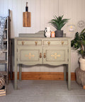 A vintage sage-green sideboard with gold accents stands against a white paneled wall, surrounded by plants, a wooden ladder with a throw, a furry stool, and rustic decor items.