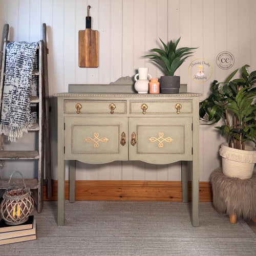A vintage sage-green sideboard with gold accents stands against a white paneled wall, surrounded by plants, a wooden ladder with a throw, a furry stool, and rustic decor items.