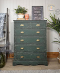 A vintage green chest of drawers with gold handles sits in a cozy room, decorated with plants, a sign reading "This is a house where love dwells," a mug, a folded blanket on a ladder, and a rug on the wooden floor.