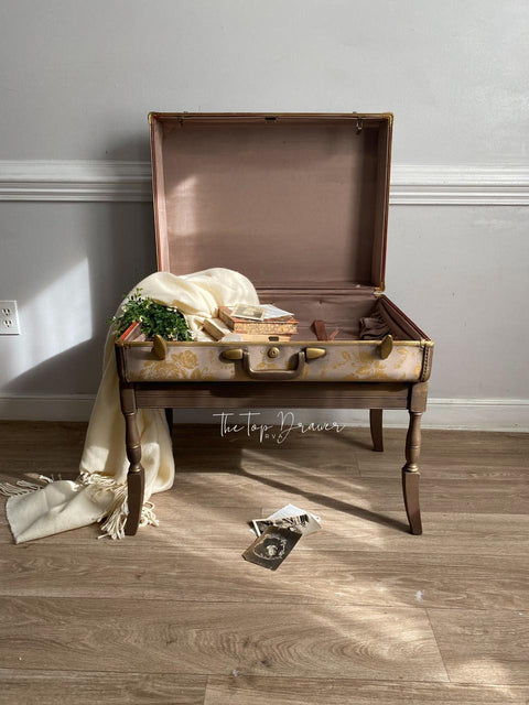 Dixie Belle Paint Chocolate Chalk Mineral Paint is the perfect paint for any DIY project!
An open vintage suitcase converted into a table, holding books, a plant, and decorative items. A cream-colored blanket drapes over one side. The setup is on a wooden floor against a gray wall with white trim.