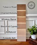 Wooden plank samples in six different shades are displayed vertically. Starting from the top: Tobacco Road, Charred Hickory, Whiskey Barrel, Bleached Cypress, AuNaturel, and Fireside Ash. A potted plant and basket are nearby.