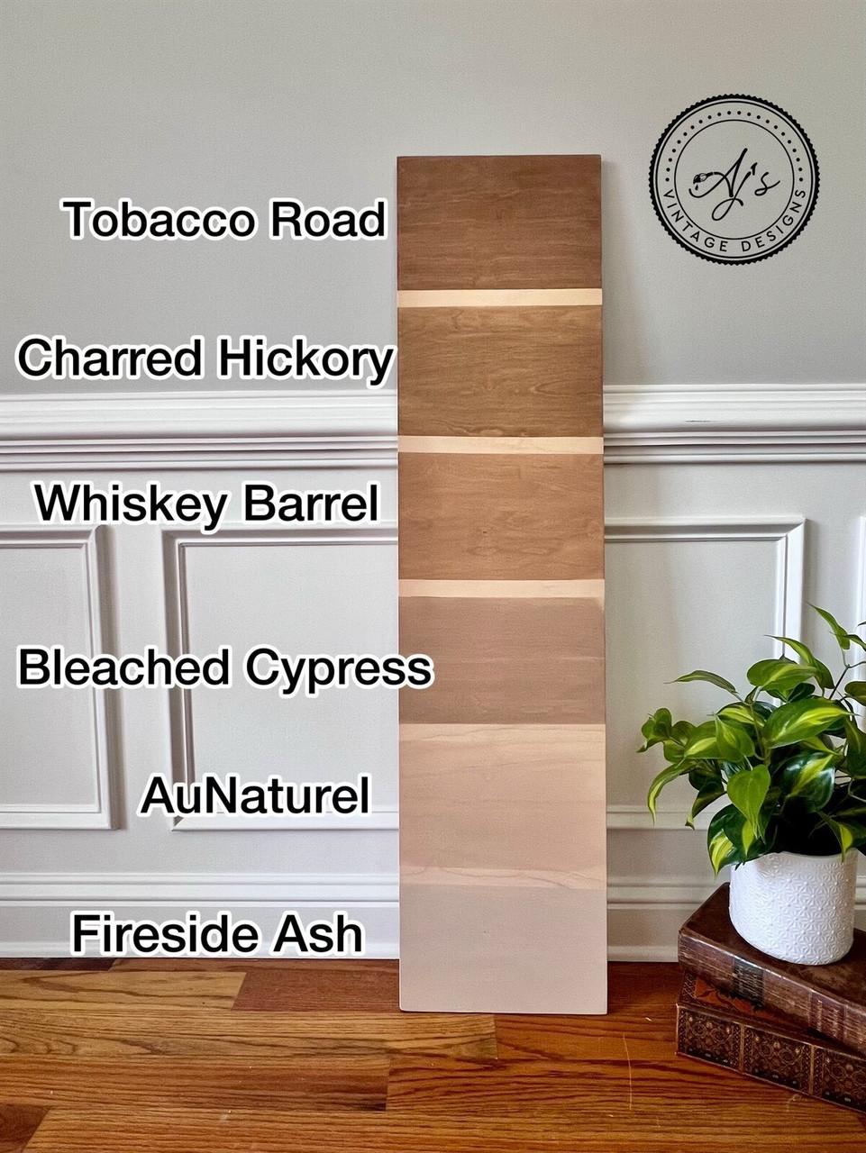 Wooden plank samples in six different shades are displayed vertically. Starting from the top: Tobacco Road, Charred Hickory, Whiskey Barrel, Bleached Cypress, AuNaturel, and Fireside Ash. A potted plant and basket are nearby.