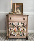 An ornate dresser with floral designs sits against a light gray wall. It has four drawers, decorative handles, and is topped with a framed portrait, candles, and a small bird statue. Scattered papers lie on a patterned floor.