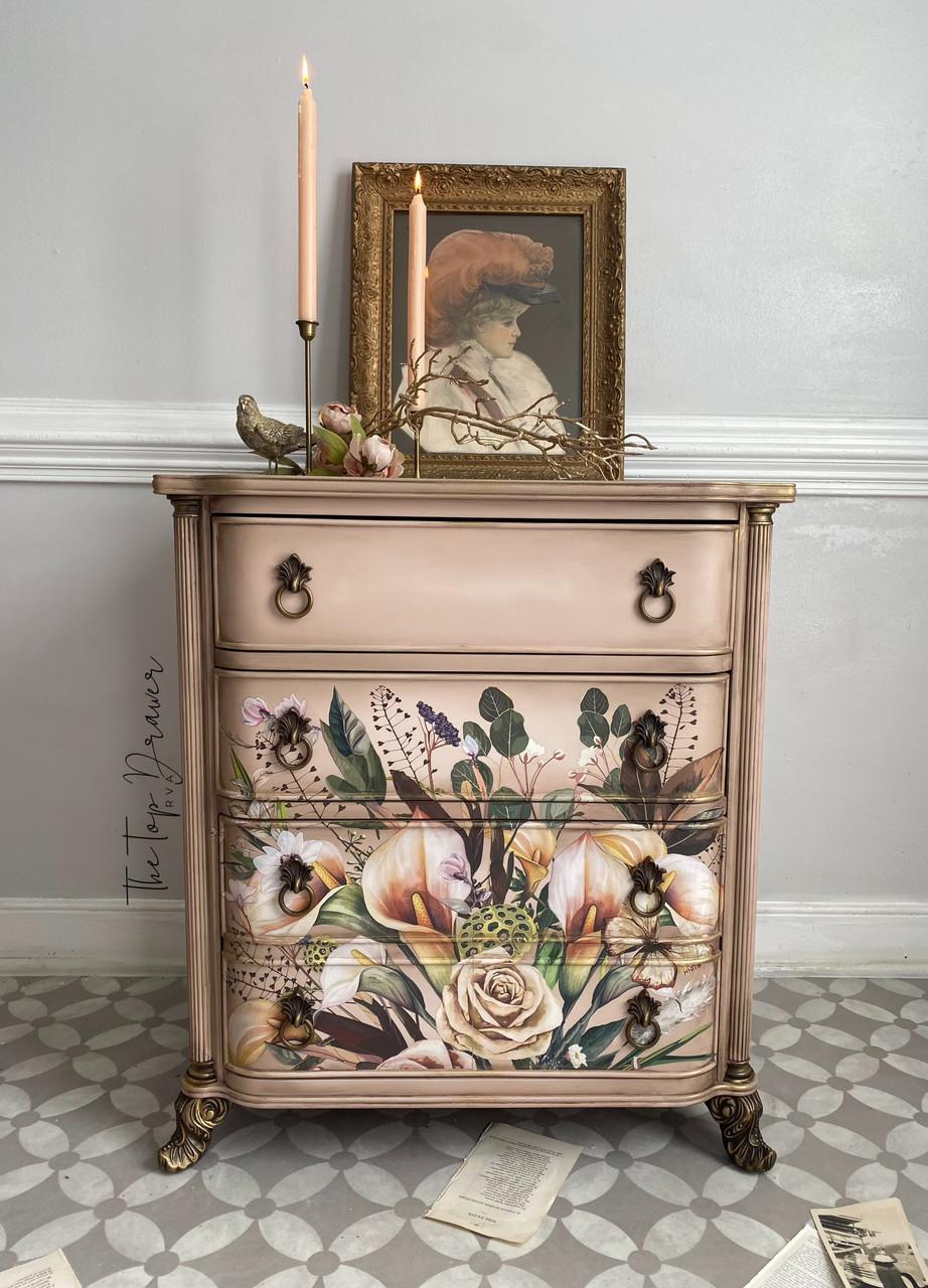 An ornate dresser with floral designs sits against a light gray wall. It has four drawers, decorative handles, and is topped with a framed portrait, candles, and a small bird statue. Scattered papers lie on a patterned floor.
