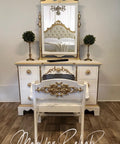 A white and gold vintage vanity with a matching chair sits on a wood floor. The vanity has ornate details, a large mirror, and two small topiary plants. A tufted bed is reflected in the mirror. Text reads, â€œMad for Rehab by Aleshia.â€.