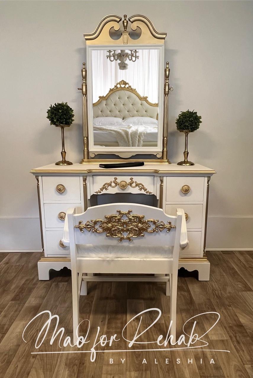 A white and gold vintage vanity with a matching chair sits on a wood floor. The vanity has ornate details, a large mirror, and two small topiary plants. A tufted bed is reflected in the mirror. Text reads, â€œMad for Rehab by Aleshia.â€.