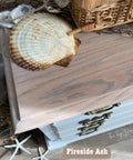 A close-up of a light wood cabinet displaying coastal decor. Sea shells, a fishing net, and a wicker basket adorn the top. A label reads "Fireside Ash" near the wood. Nautical elements like a starfish and rope are visible.