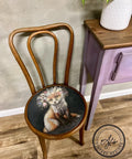 A wooden chair with a circular seat cushion featuring a painted fox wearing a floral crown. The chair is positioned on a wooden floor beside a small purple-painted table with a potted plant on top. The wall behind is textured. An "AJ's Vintage Designs" watermark logo is on the floor.
