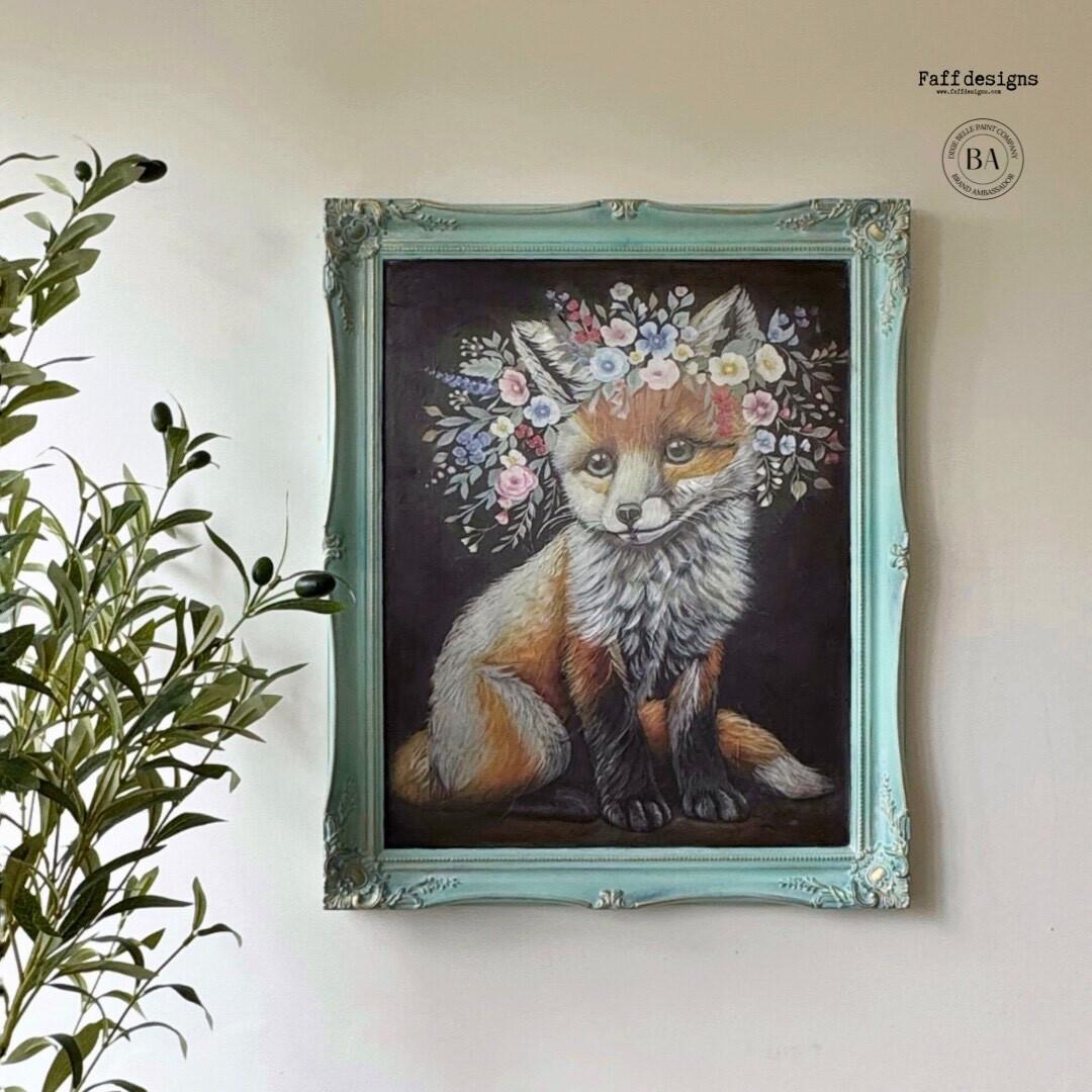 A decorative painting of a fox with a crown of colorful flowers is displayed in an ornate turquoise frame. The artwork is mounted on a white wall, with a leafy green plant placed to the left of the painting.