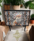 A painted dresser stands in a room adorned with lush green plants. The dresser features an intricate design of a fox wearing a flower crown, framed by an ornate black and white pattern. The room includes beige armchairs and a rug with the logo "TLC Vintages."