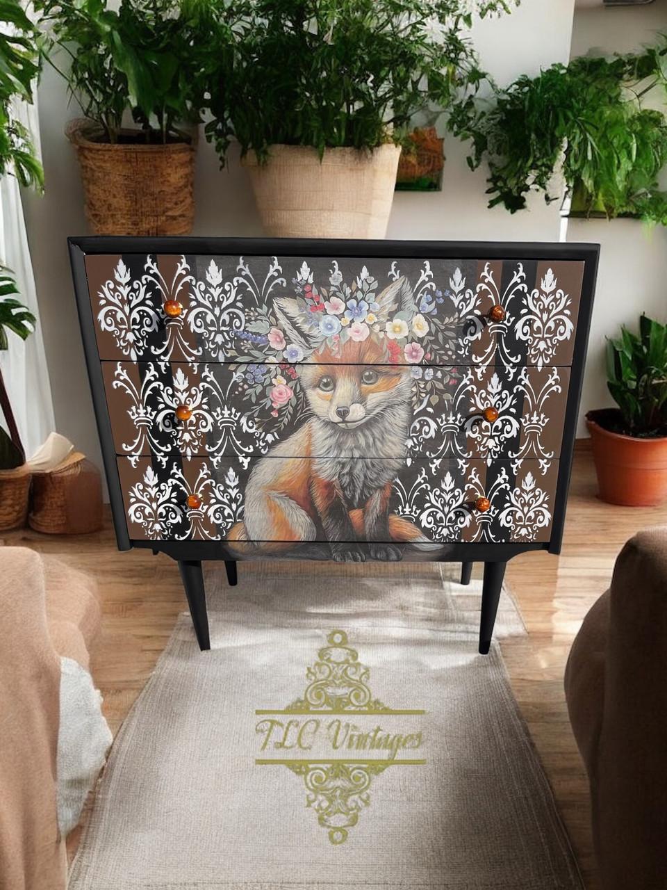 A painted dresser stands in a room adorned with lush green plants. The dresser features an intricate design of a fox wearing a flower crown, framed by an ornate black and white pattern. The room includes beige armchairs and a rug with the logo "TLC Vintages."