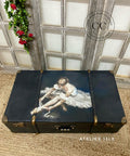 A suitcase with a painting of a seated ballerina adjusting her ballet slippers on the lid. The chest is placed on a woven rug in front of a decorative window pane with red flowers. The logos "CC/ Dixie Belle Paint Company Content Creator" and "Atelier 13LX" are visible near the chest.