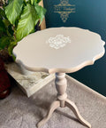 A small, round, light beige vintage side table with scalloped edges and white floral detailing on top stands on a three-legged pedestal base next to a green leafy plant on a carpeted floor.