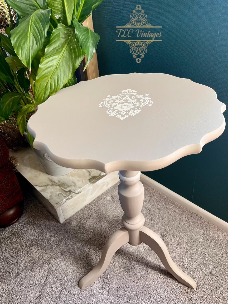A small, round, light beige vintage side table with scalloped edges and white floral detailing on top stands on a three-legged pedestal base next to a green leafy plant on a carpeted floor.