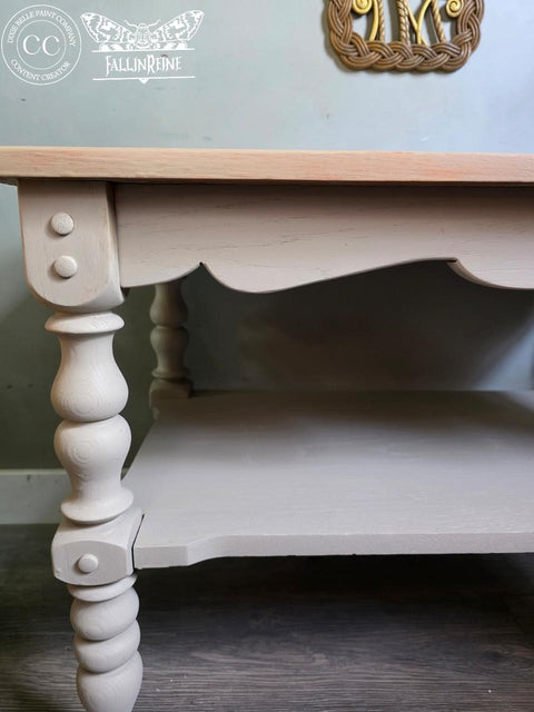 A close-up of a light beige wooden table with ornate, turned legs and a scalloped apron. The table sits on a dark wood floor, and logos are visible in the top left corner of the image.