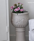 A tall, ornate beige planter with pink flowers sits next to a white locker and a white decorative statue. The planter features embossed floral designs. "Sisterhood Furniture" watermark appears in the top right corner.