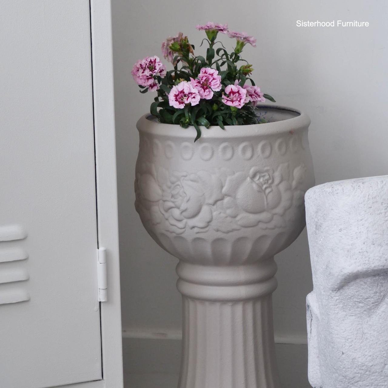 A tall, ornate beige planter with pink flowers sits next to a white locker and a white decorative statue. The planter features embossed floral designs. "Sisterhood Furniture" watermark appears in the top right corner.