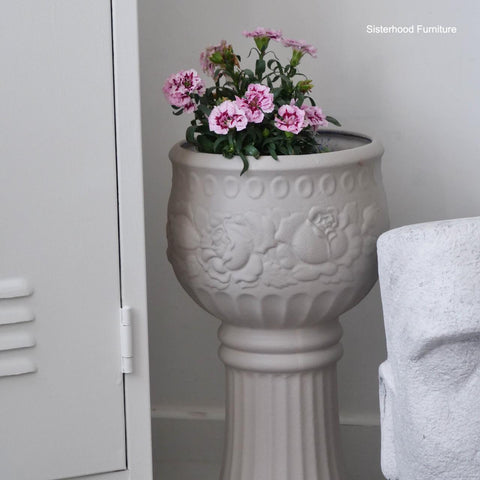 A tall, ornate beige planter with pink flowers sits next to a white locker and a white decorative statue. The planter features embossed floral designs. "Sisterhood Furniture" watermark appears in the top right corner.