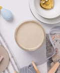 A top-down view of a jar filled with beige paint, surrounded by a decorative plate with a gold heart, ribbon spools, a blue perfume bottle, a small brush, and patterned fabric on a white surface.
