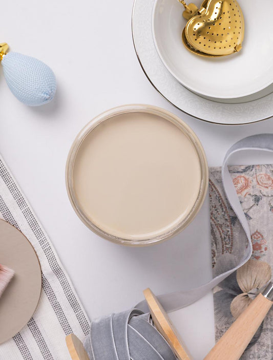 A top-down view of a jar filled with beige paint, surrounded by a decorative plate with a gold heart, ribbon spools, a blue perfume bottle, a small brush, and patterned fabric on a white surface.