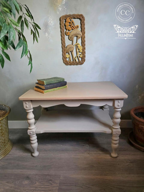 A painted wooden table with ornate legs stands on a gray floor. Three stacked vintage books sit on top. A decorative framed artwork hangs on the wall above, near potted plants.