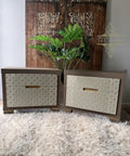 Two modern nightstands with geometric, textured fronts and inset handles sit side by side on a fluffy beige rug. Green plants and a carved wooden door serve as the background decor.