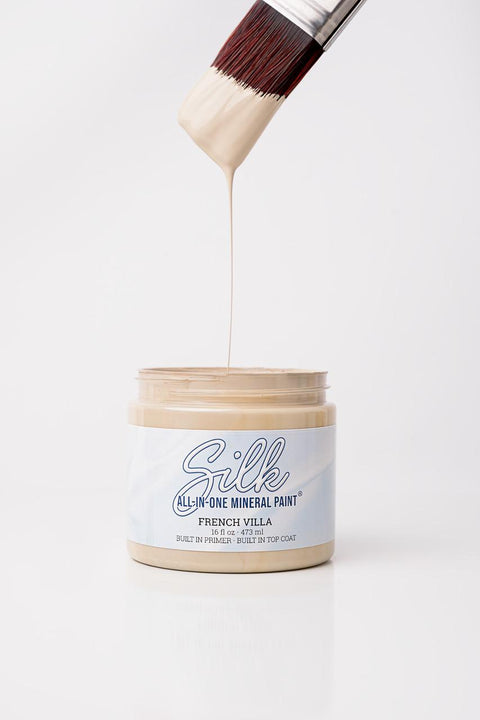 A paintbrush dripping beige paint above an open jar labeled "Silk All-in-One Mineral Paint, French Villa" against a plain white background.
