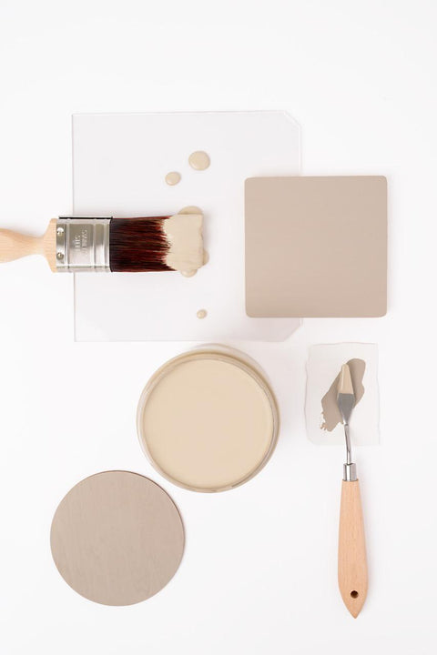 A flat lay of neutral-colored paint samples, a round paint can, a paintbrush, a palette knife, and paint swatches arranged neatly on a white background.