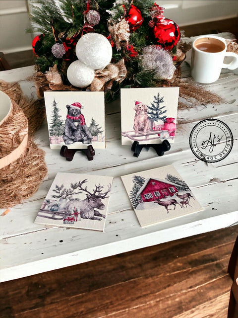 A table displays festive holiday coasters with the Frosty Forest Friends Decoupage Paper. Each coaster features a winter scene: a dog in a red Santa hat, a bear in a red scarf and hat next to gifts, a moose in a snowy field, and a red cabin with a family of deer in front. A centerpiece with ornaments, a cup of coffee, and rustic decor accents the setting.