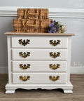 A small white dresser with four drawers featuring ornate brass handles stands against a gray wall. On top, there are three woven baskets and a bouquet of purple and white flowers. The floor is wooden.