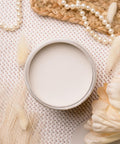 Open jar of cream-colored paint on a textured surface, surrounded by decorative elements like pearl necklaces, dried flowers, and woven material. The setup conveys a soft, elegant, and natural aesthetic.