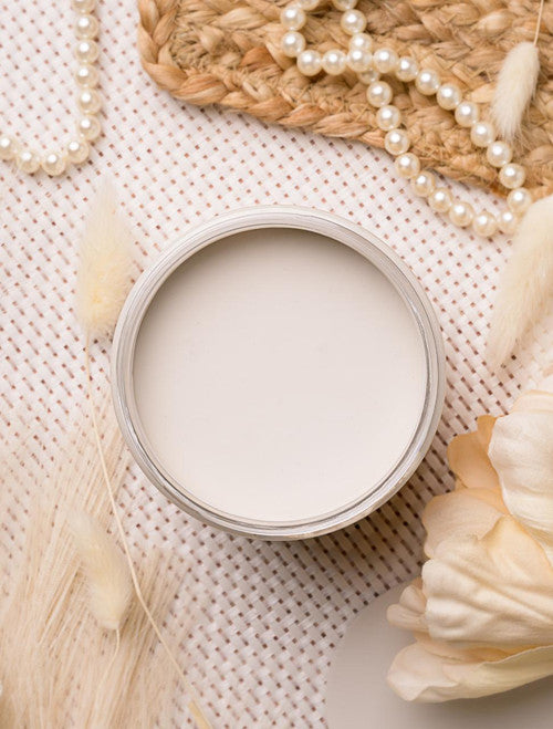 Open jar of cream-colored paint on a textured surface, surrounded by decorative elements like pearl necklaces, dried flowers, and woven material. The setup conveys a soft, elegant, and natural aesthetic.