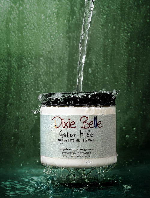 A jar of Dixie Belle Gator Hide is splashed with a stream of water against a green background, demonstrating its water-repellent properties. Water droplets are seen around and on the container.