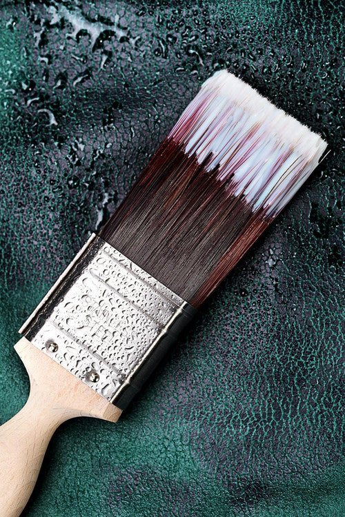 A close-up of a paintbrush with a wooden handle resting on a dark green textured surface with water droplets. The brush bristles are dipped in white topcoat.