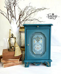 Blue painted end table with gold knobs, blended with white in the center front of the cabinet door. The Gilded Damask Transfer has been applied on top of the white blending on the insert of the cabinet face. Staged next to a stack of wooden boxes with a gold crown on top.