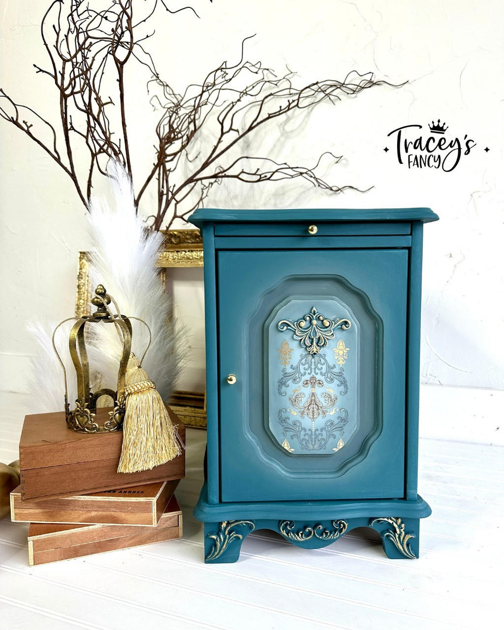 Blue painted end table with gold knobs, blended with white in the center front of the cabinet door. The Gilded Damask Transfer has been applied on top of the white blending on the insert of the cabinet face. Staged next to a stack of wooden boxes with a gold crown on top.