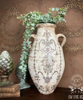 Large white distressed concrete vase with 2 handles and faux eucalyptus overflowing out of it. The Gilded Damask Transfer has been applied to the front. Staged next to a stack of books with a  patina'd sculpture of a pinecone on top.